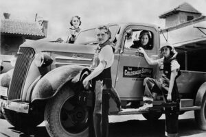 An archival 1944 black and white photograph of women in overalls posing near a Colorado Supply Division pickup truck.