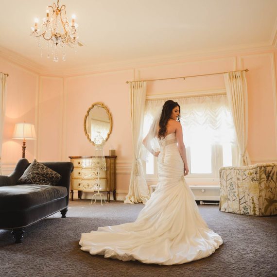 Bride poses in the middle of a light filled, pink dressing suite decorated with beautiful furniture, mirrors and a crystal chandelier.
