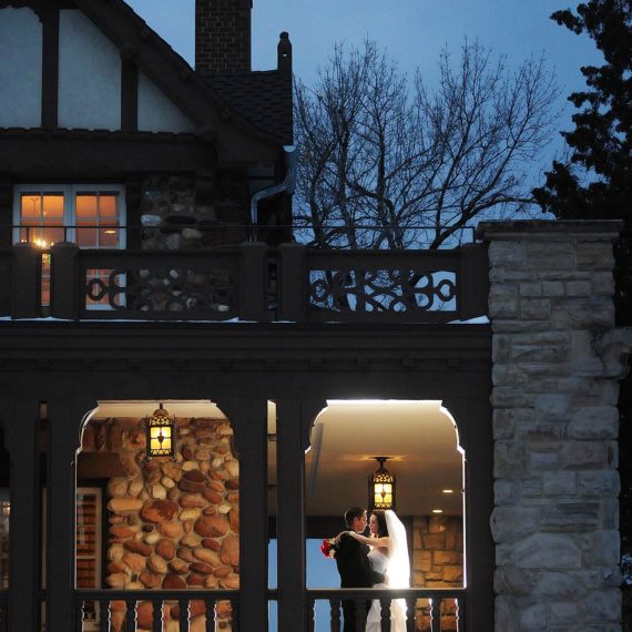 A couple embraces on the lower porch of the Highlands Ranch Mansion under a dusky sky with light snow gracing the deck and porch railings.