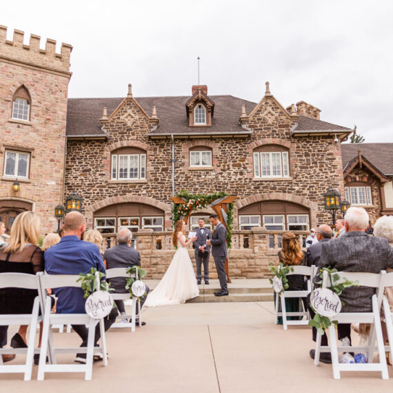 A couple exchanges vows on the front steps of a castle-like stone and tudor Mansion. They are framed by a flower covered arch, and their guests sit in white chairs in the foreground.