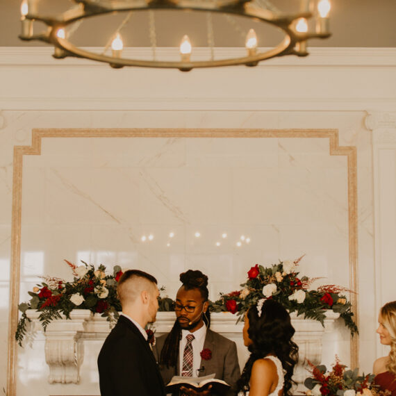 A couple holds hands during their wedding ceremony and listens to the officiant as they all stand in front of a fireplace draped in flowers under an antique metal chandelier.