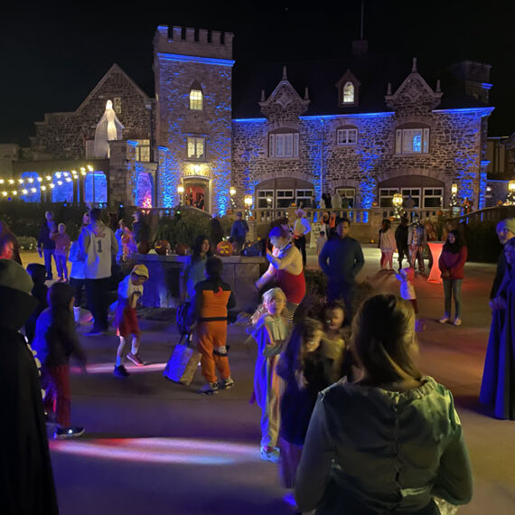 Adults and kids in costumes dance and mill around the front of the Highlands Ranch Mansion on Halloween. The castle-like stone building in the background is lit up with blue and orange lights lending a spooky ambiance.