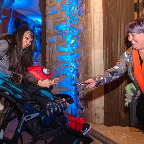 An adult in costume smiles while her toddler sitting in a stroller takes a candy bar from two women passing out candy for Halloween.