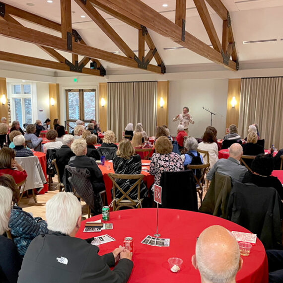 Landmark Lecture guests sit at tables covered in red tablecloths under the wooden beams of the Great Hall while listening to a local author presenting from the front of the room.