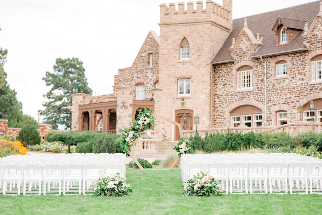 Rows of white chairs are set for a ceremony on the lawn in front of a castlelike Mansion.
