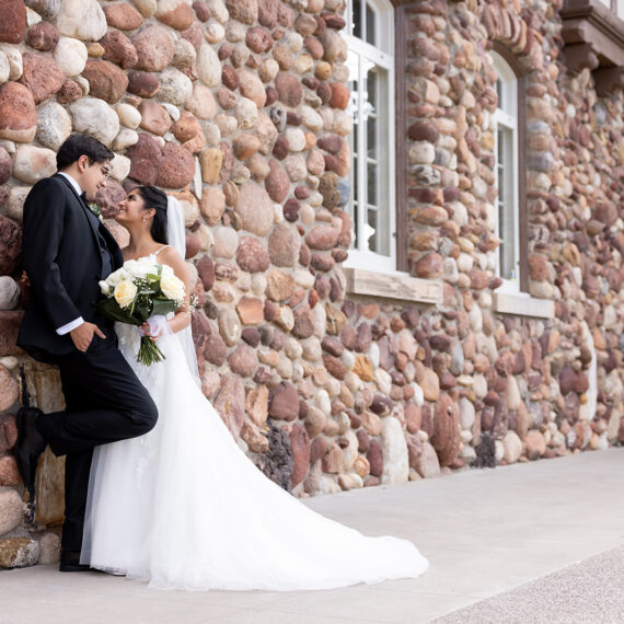 A bride and groom pose for a photo in front of the stone wall of the Mansion.