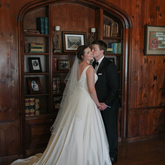 A bride and groom share a kiss in front of the Library bookcase.